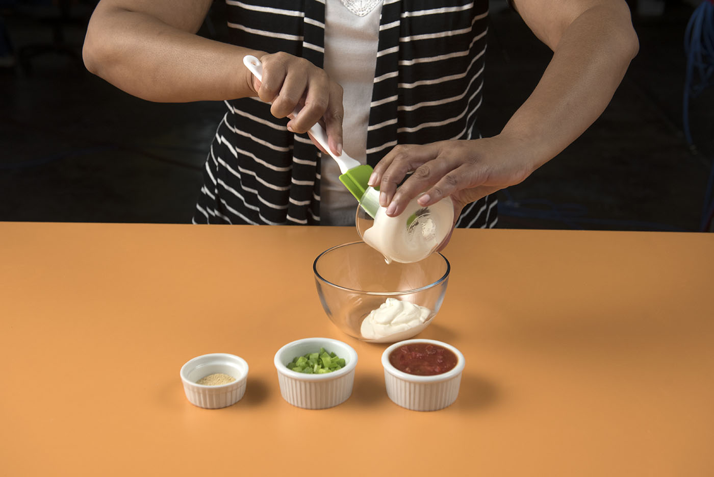 A Woman prepares to mix mayonaise, salsa, green onions, and spices together in a glass bow.