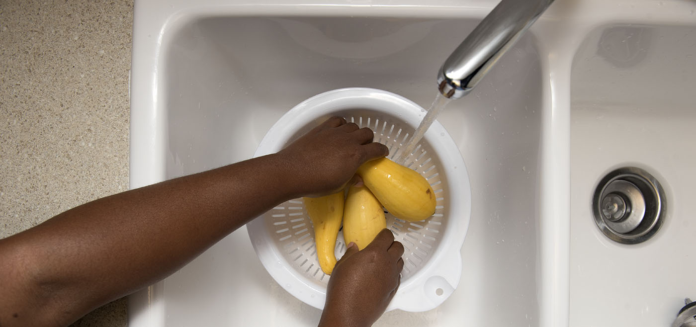 A woman's hands are shown washing three squash in a colander as water runs over them. 
