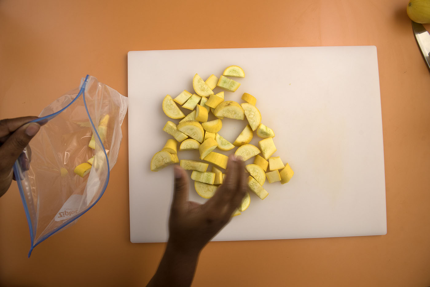 A woman moving cut yellow squash from the cutting board to a storage bag.