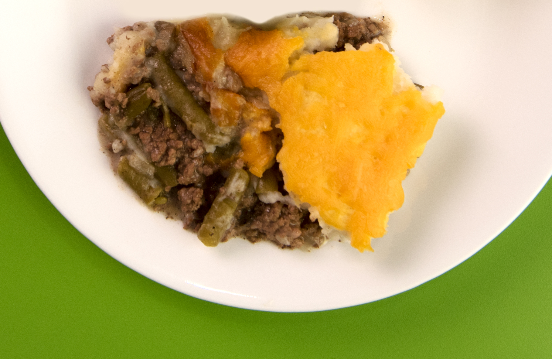 A white plate filled with Shepherd's Pie; a recipe using ground beef, green beans, mashed potatoes, and cheese.