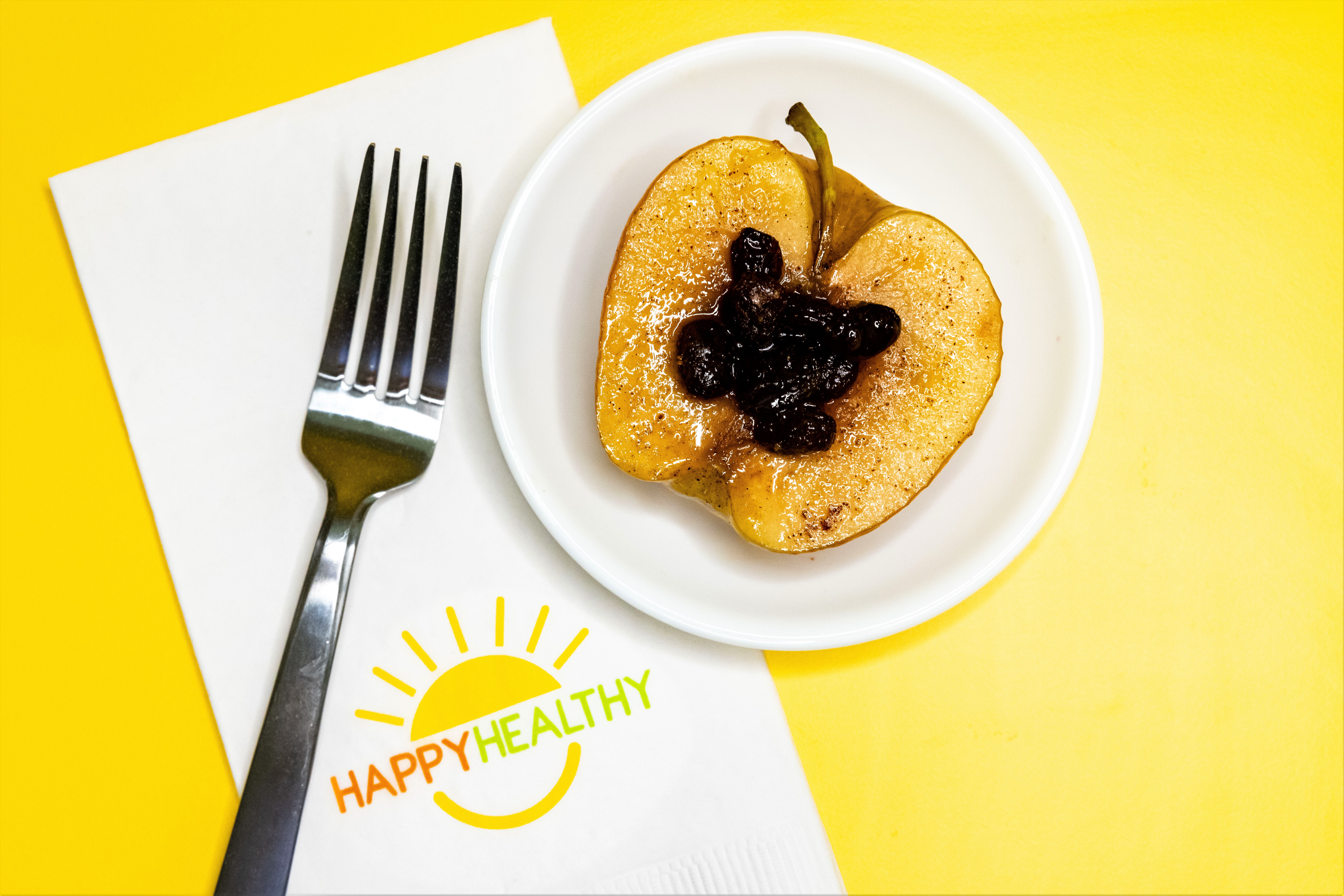 Microwaved apple half on a white plate with raisins on top. HappyHealthy napkin and fork to left of plate.
