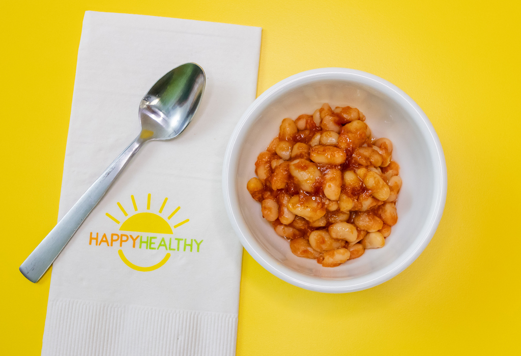 A white bowl with baked beans, next to a HappyHealthy napkin and a spoon, all on a yellow background