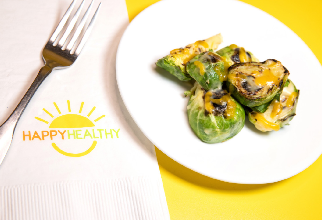 Baked Brussels Sprouts on white plate next to HappyHealthy napkin and fork