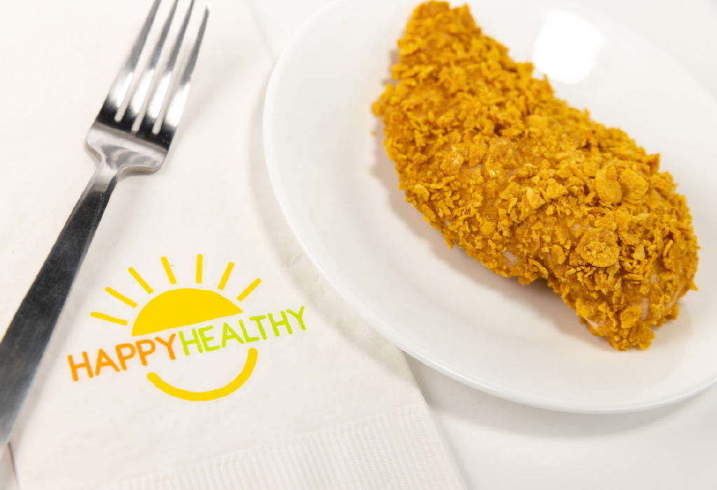 A baked flaked chicken breast on a white plate next to HappyHealthy napkin and fork