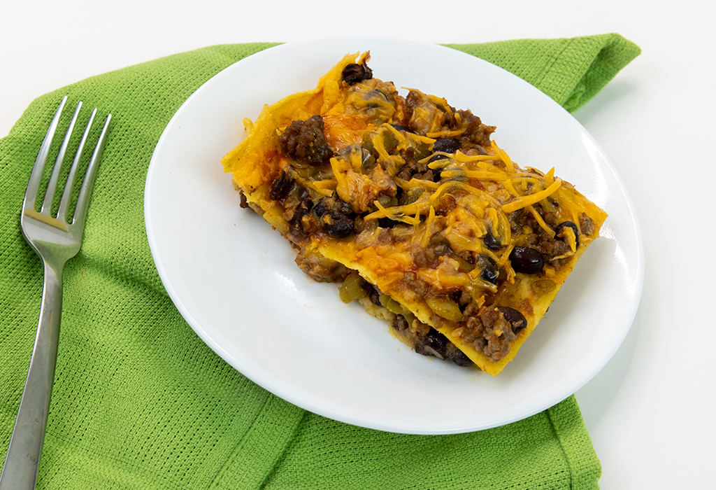 A slice of beef enchilada casserole on a white plate laying on a green towel