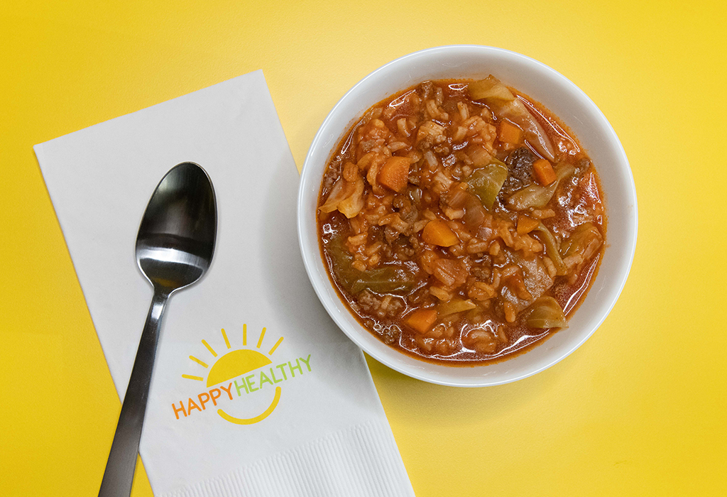 A bowl of cabbage roll soup next to a spoon and HappyHealthy napkin