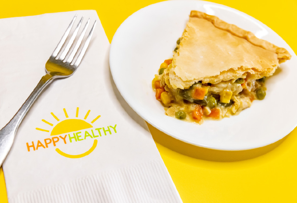A slice of chicken pot pie on a white plate next to a fork and HappyHealthy napkin