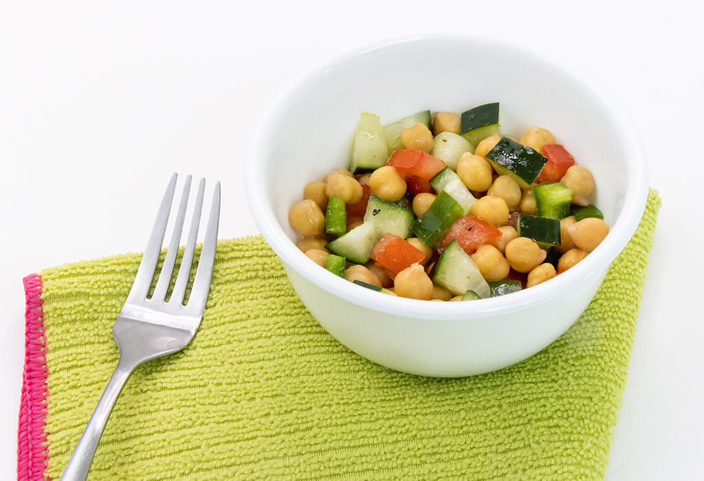 A white bowl filled with squash, tomatoes, and zucchini next to a fork and green cloth napkin
