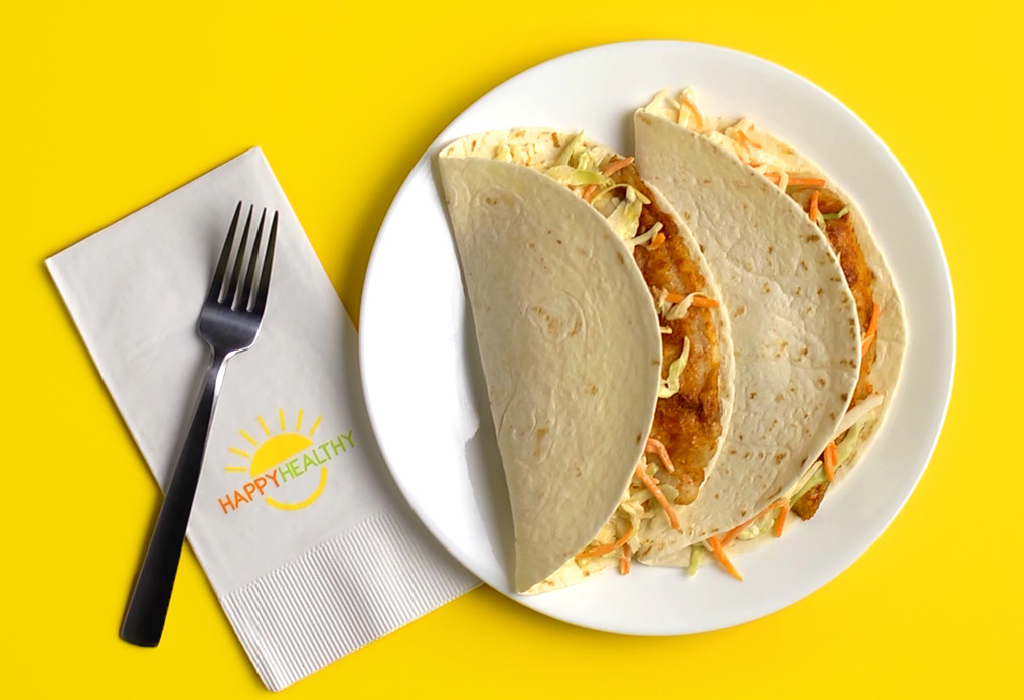 A white plate with two fish tacos next to a fork and Happy Healthy napkin