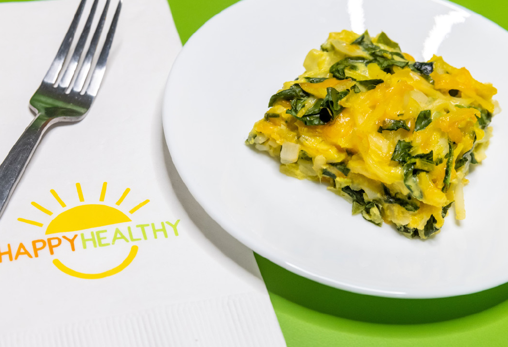 A square piece of hash brown casserole on a white plate next to a HappyHealthy Napkin and fork