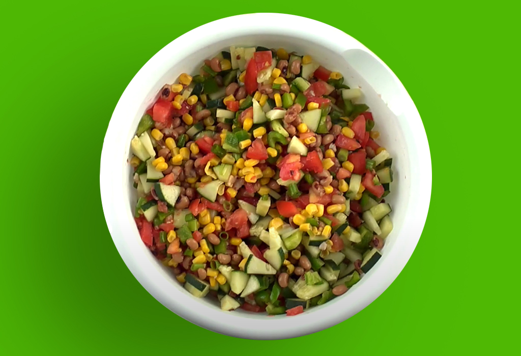 White bowl of chopped vegetables (corn, tomato, cucumber, etc) with green background.