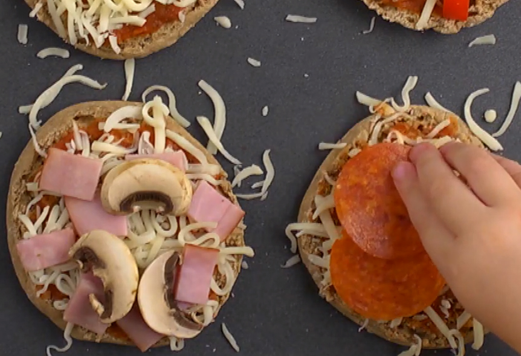 Several mini pizzas on a tray with a hand topping one with pepperoni