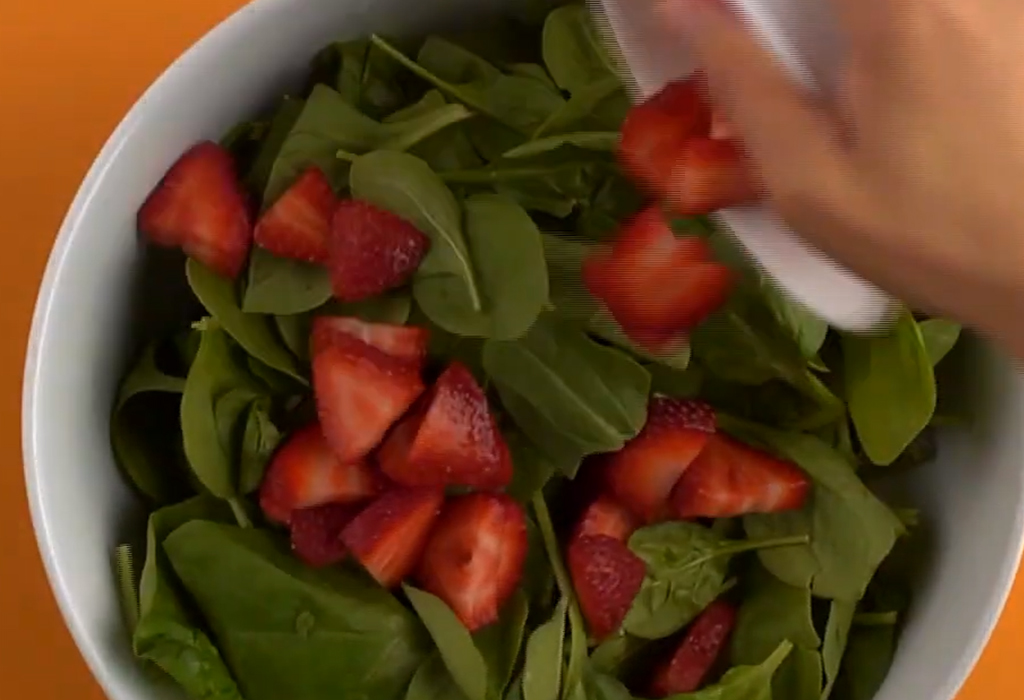 A hand adding chopped strawberries to a bowl of spinach