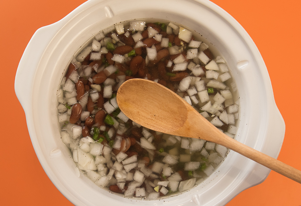 Wooden spoon positioned over a slow cooker of beans, garlic, jalapenos, and onions
