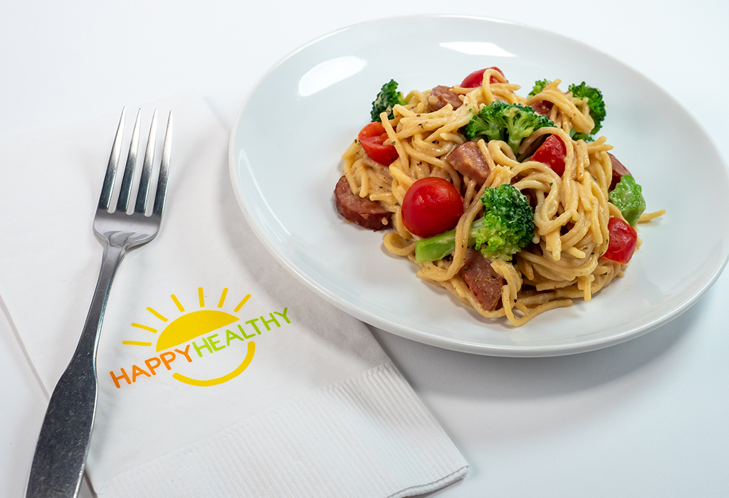 A small bowl of pasta with broccoli, ham and tomatoes  next to a Happy Healthy napkin and fork