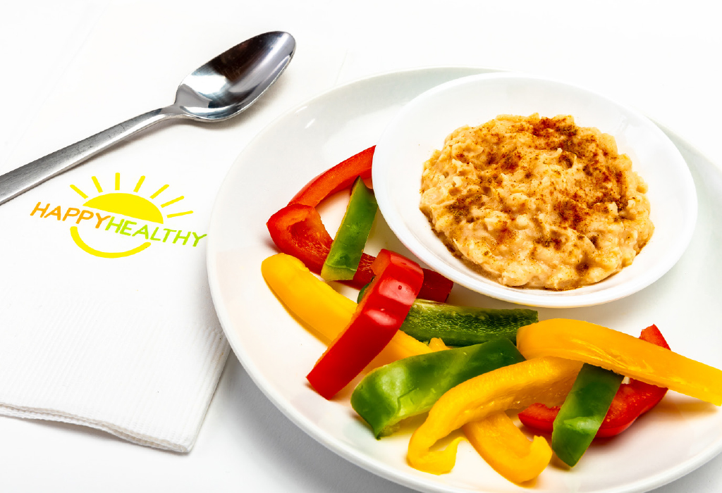 Bowl of hummus on white plate with spread of different colored peppers with HappyHealthy napkin and spoon to the side.