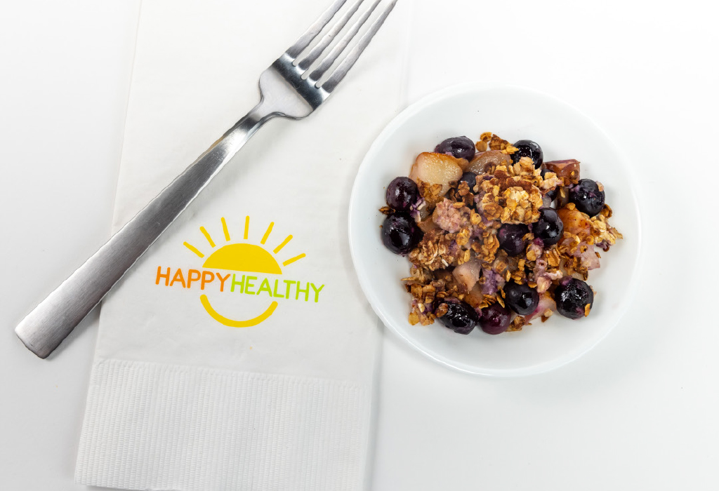 A bowl of pear and blueberry crisp next to a fork and HappyHealthy Napkin