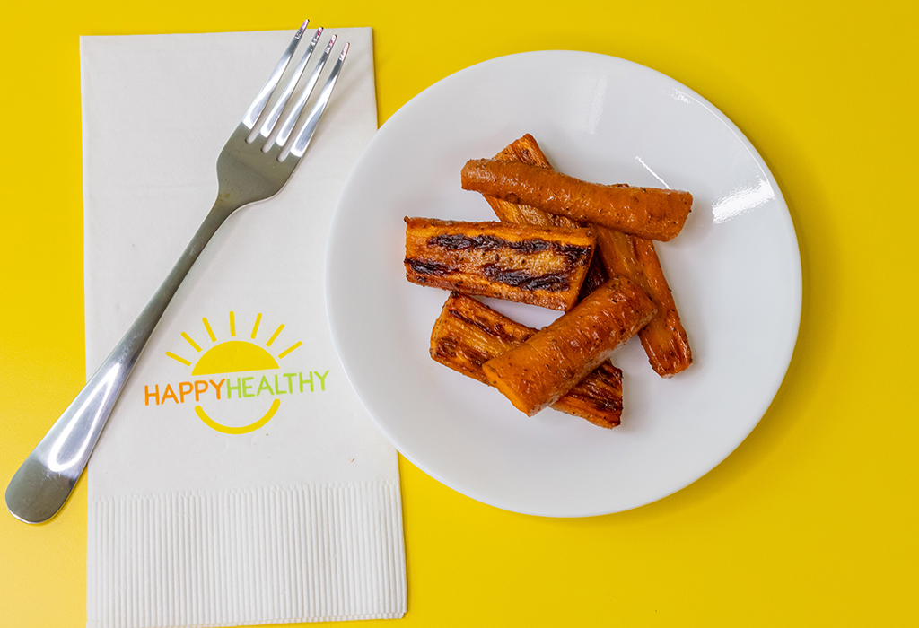 A white plate with roasted carrots next to a fork and HappyHealthy napkin on a yellow background