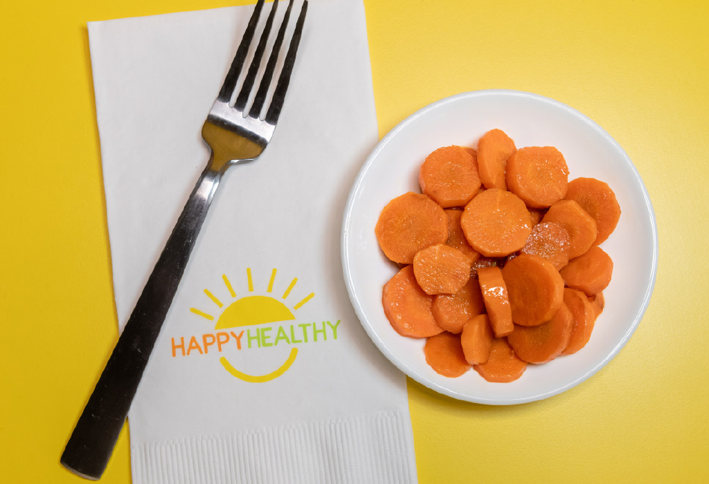 A white bowl of sautéed carrots next to a fork and HappyHealthy napkin