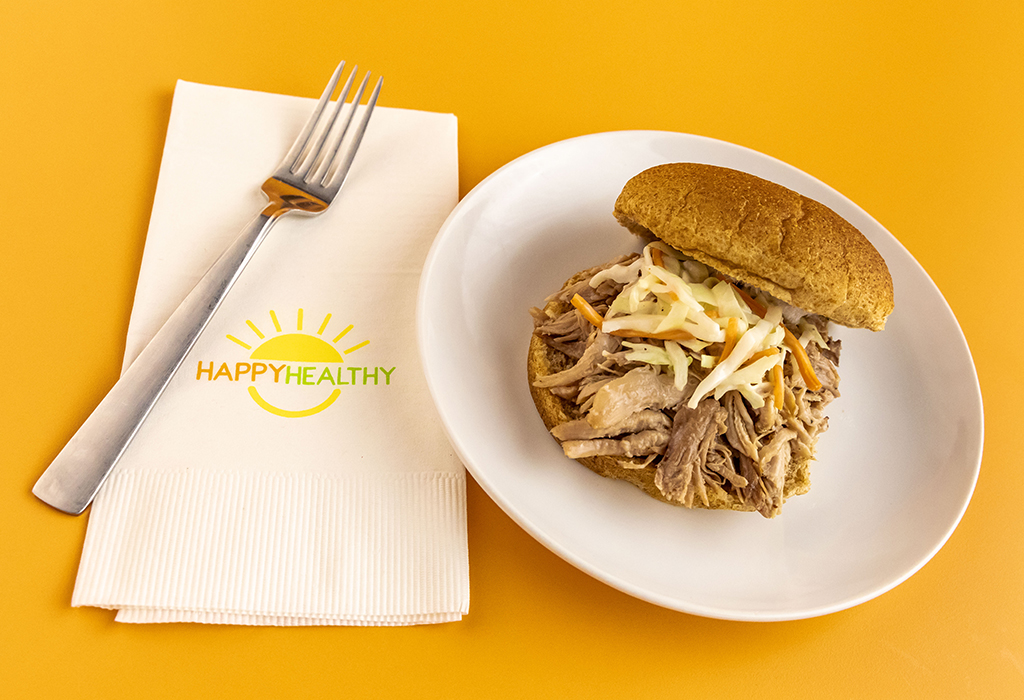 Slow Cooker Pulled Pork on a bun, plated on a white plate next to a HappyHealthy napkin and fork