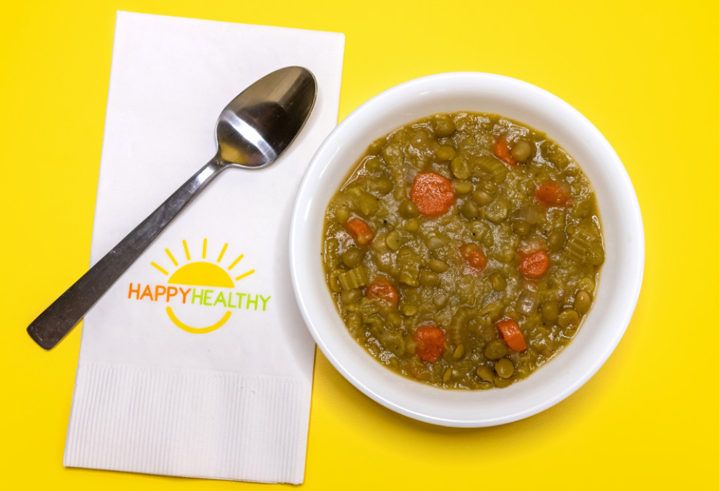 A bowl of split pea soup in a white bowl next to a HappyHealthy napkin and spoon