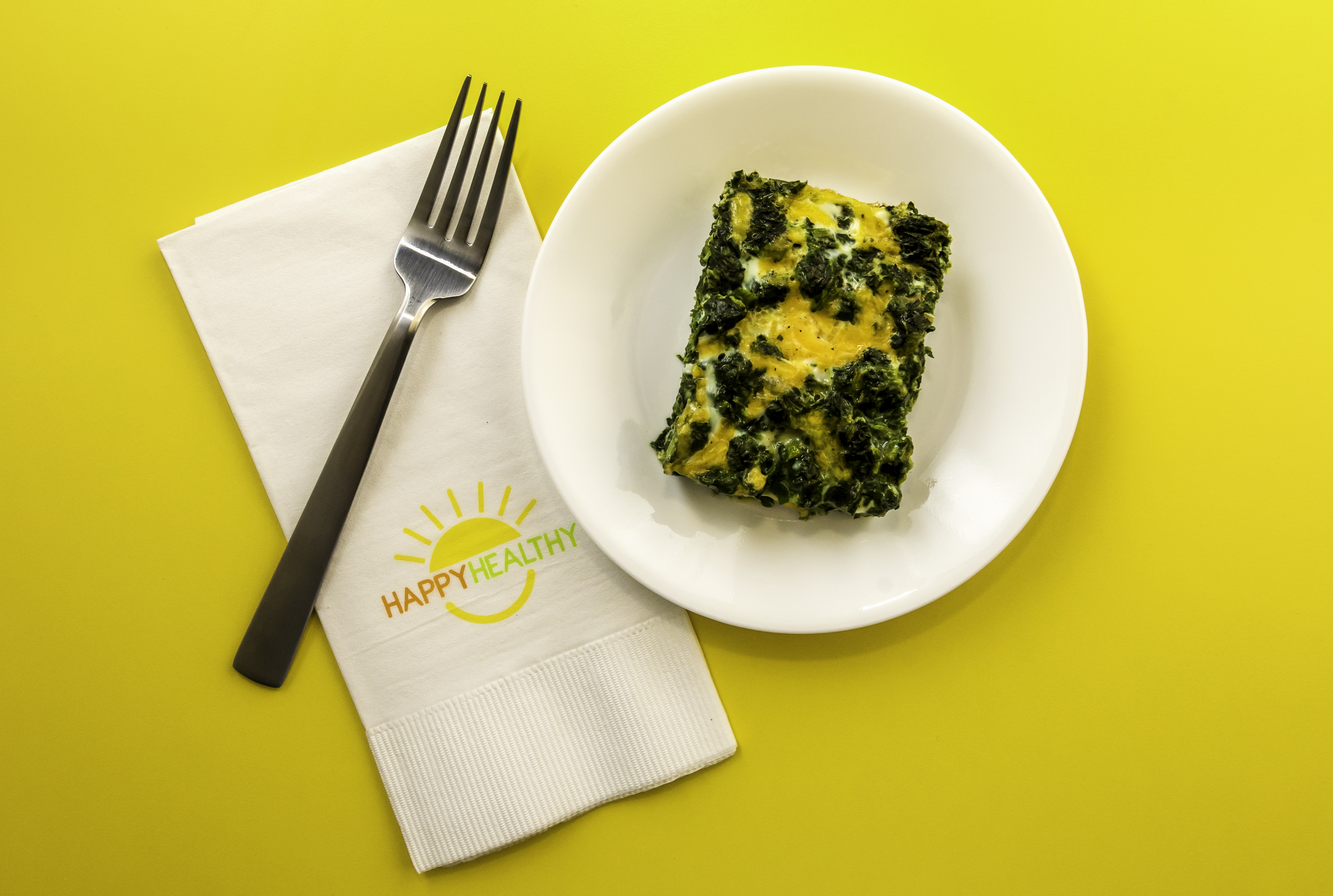 A serving of egg and spinach casserole on a white plate with a fork and napkin to the side.