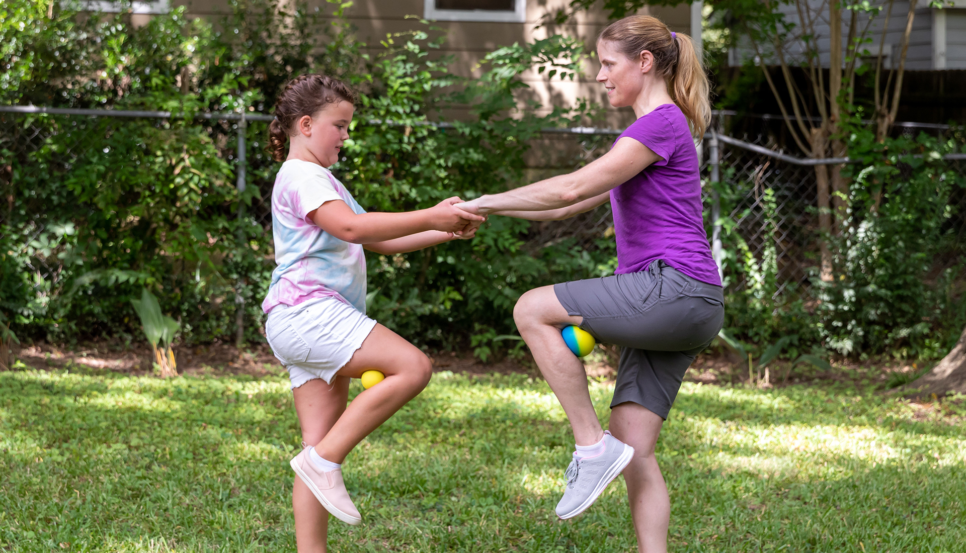 A mother and daughter outside holding hands and each balancing on one foot