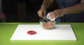 A woman places seasonings in a clear bowl on a white cutting board to prepare the all purpose seasoning.
