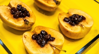 Four baked apples halves with raisins on top of each in a square clear glass baking dish.