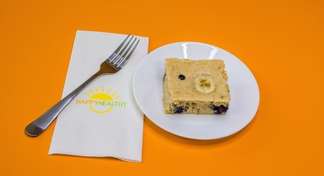 A small white plate with a square of blueberry and banana sheet pan pancakes next to a fork and happyhealthy napkin