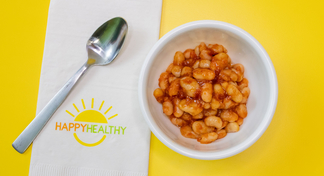 A white bowl with baked beans, next to a HappyHealthy napkin and a spoon, all on a yellow background