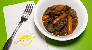 A white bowl with beef stew next to a spoon and HappyHealthy napkin