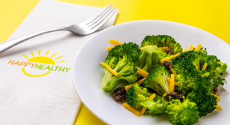 Broccoli salad on white plate next to white HappyHealthy napkin and fork.