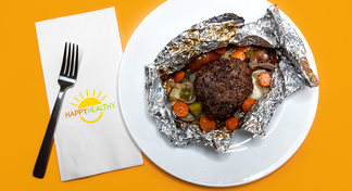 An open foil packet with a patty, cooked potatoes, peppers and carrots on a white plate next to a fork and HappyHealthy napkin