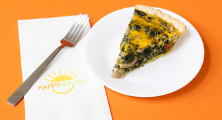 A slice of vegetable quiche on a white plate next to a fork and Happy Healthy napkin