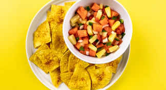 A white bowl of watermelon salsa on a white plate of tortilla chips