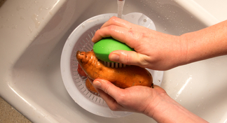 Two hands scrubbing potato with a brush in a sink with running water over a colander 
