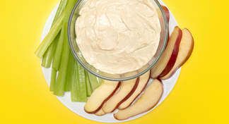 A bowl of peanut butter dip surrounded by celery sticks and apple slices on a white plate
