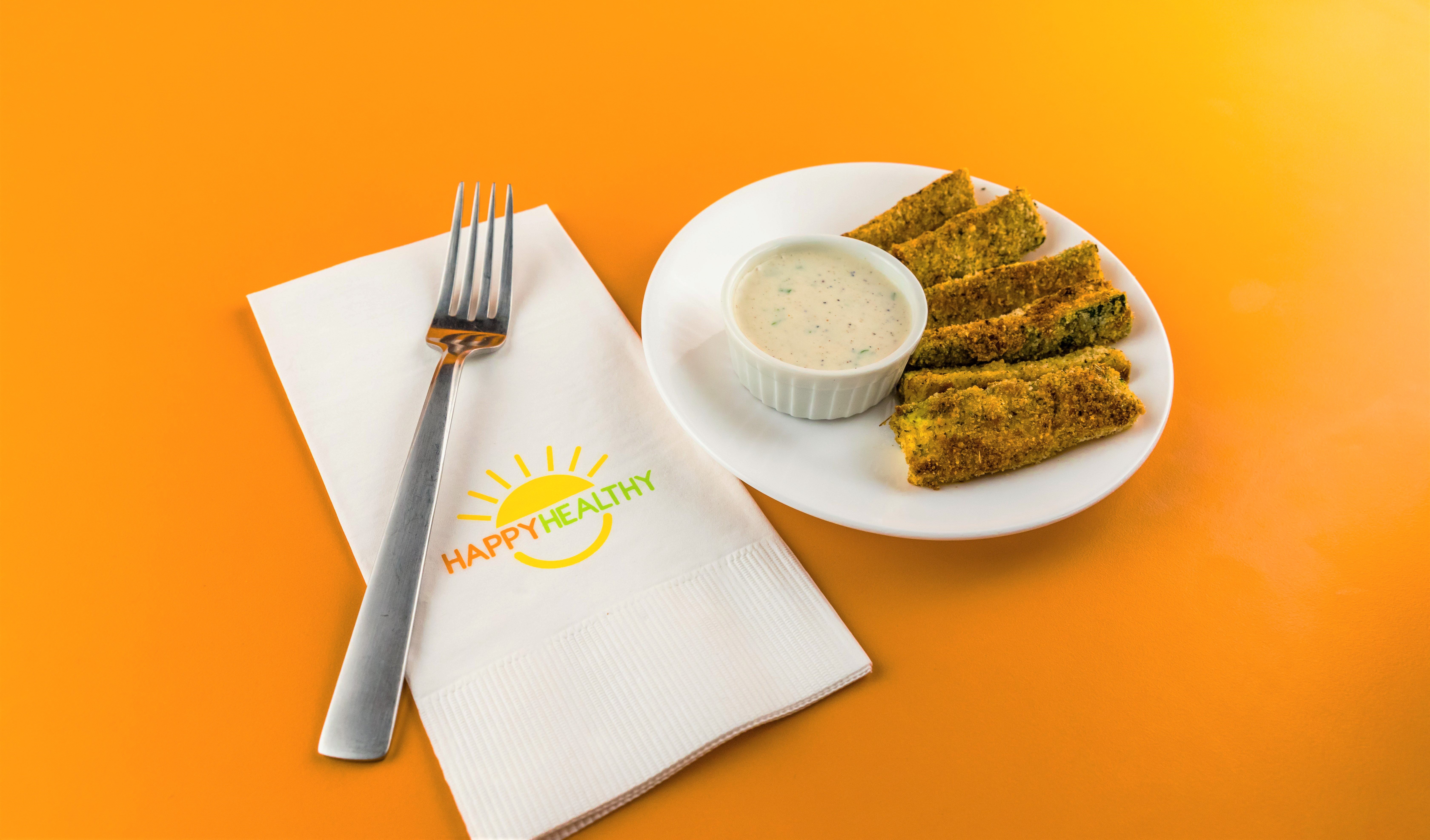 A plate of baked zucchini and ranch dipping sauce next to a plate and HappyHealthy napkin