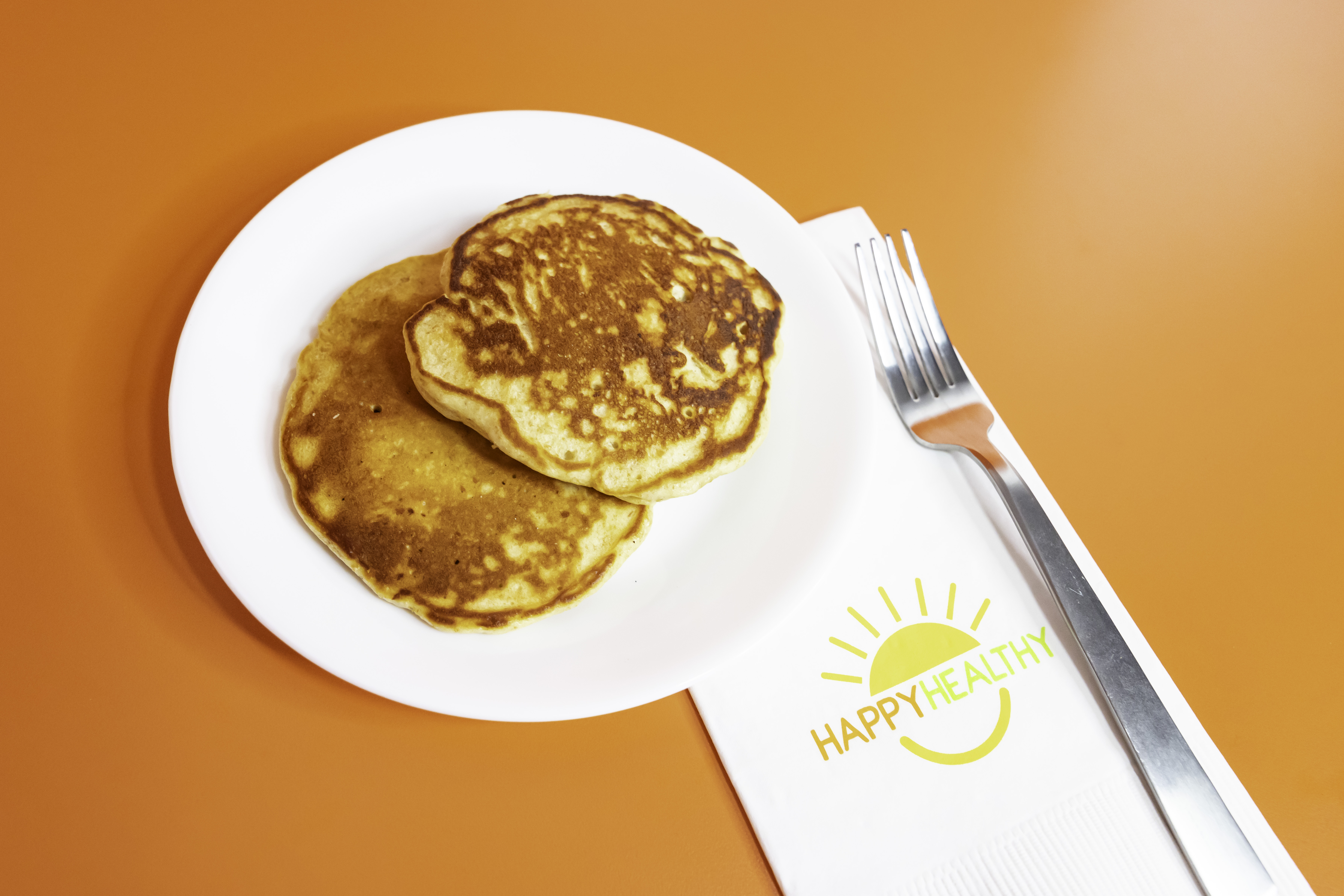 A white plate with two pancakes on an orange background, next to a napkin and fork
