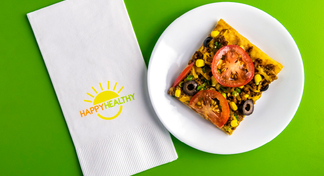 Fork and napkin with plate with a square pizza slice covered with tomatoes, olives, onions, beef, corn, and cheese