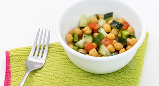 A white bowl filled with squash, tomatoes, and zucchini next to a fork and green cloth napkin