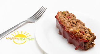 A slice of meatloaf on a white plate next to a fork and Happy Healthy napkin