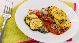 A white bowl filled with squash, tomatoes, and zucchini next to a fork and green cloth napkin